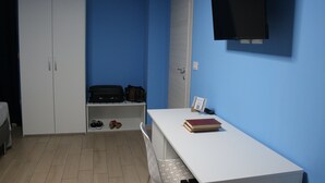 Ein Zimmer mit weißem Schreibtisch, einem Bücherregal, einem Kleiderschrank und einem an der Wand befestigten Fernseher.