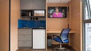 Een kleine kantoorruimte met een kitchenette, een bureau met stoel, en een aan de muur bevestigde televisie.