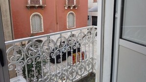 Un balcone con ringhiere in metallo bianco decorate, un edificio rosso con due finestre e balconi, e una bicicletta parcheggiata.