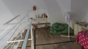Una stanza con soffitto spiovente, un letto con coperta verde, una ventola da terra e un tavolino con tovaglia rossa.
