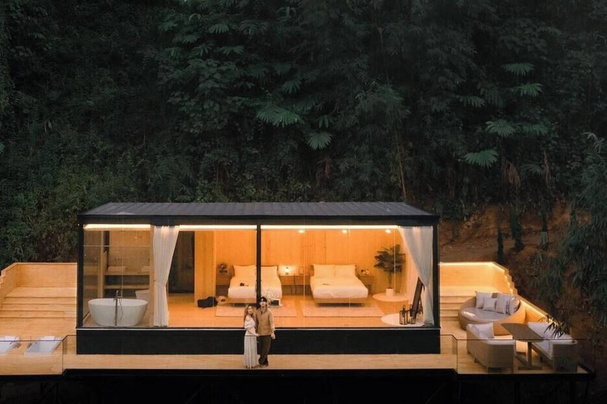 Une cabane moderne, entièrement vitrée, dotée d’une terrasse en bois, d’une salle de bain avec un lavabo et des toilettes, et d’un espace de vie comprenant un canapé et une table basse.
