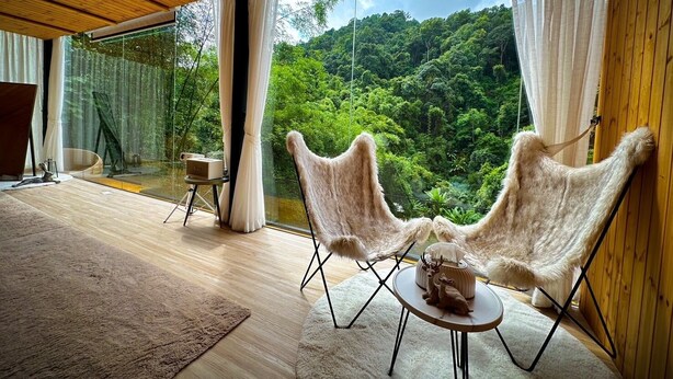 A room with a large window overlooking a forest, two fur-covered chairs, a small table with a deer figurine, and a wooden ceiling. - Morning Star Glamping (Mae On)