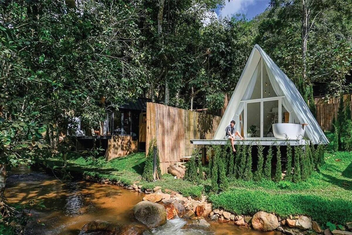 Une maison moderne avec un toit triangulaire, une petite terrasse et un ruisseau en arrière-plan.