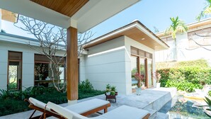 A modern outdoor lounge area with two wooden lounge chairs, a small table with potted plants, and a clear view of a pool and greenery. - Enjoy A Luxurious 5 Bedroom Villa (Da Nang)