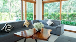 A modern living room with a grey sofa, wooden coffee table, and large windows with a view of greenery. - Enjoy A Luxurious 5 Bedroom Villa (Da Nang)