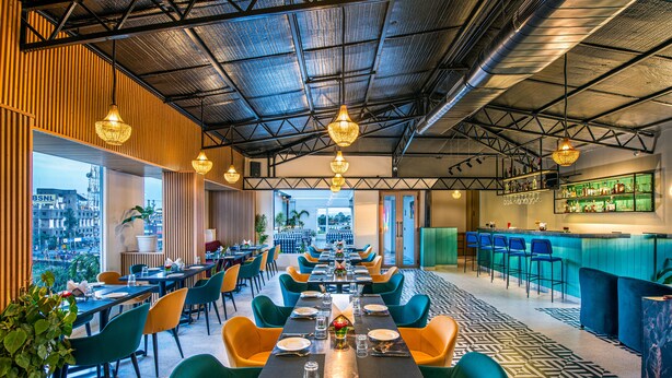 Ein modernes Restaurant mit einem langen Essbereich, mehreren Tischen und Stühlen. Es gibt eine Bar mit einer Theke und Hockern. Die Decke weist ein Metallgitter-Design auf, und es hängen Lampen ab.