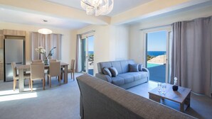 Habitación de hotel moderna con sofá gris, mesa de comedor con sillas, candelabro y vista al mar.
