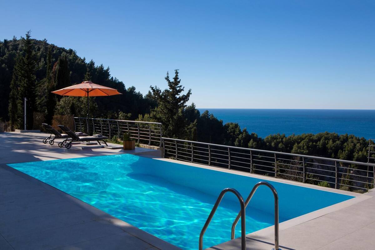 A swimming pool with clear blue water, surrounded by a metal railing, and overlooking a scenic view of the sea and forested hills.