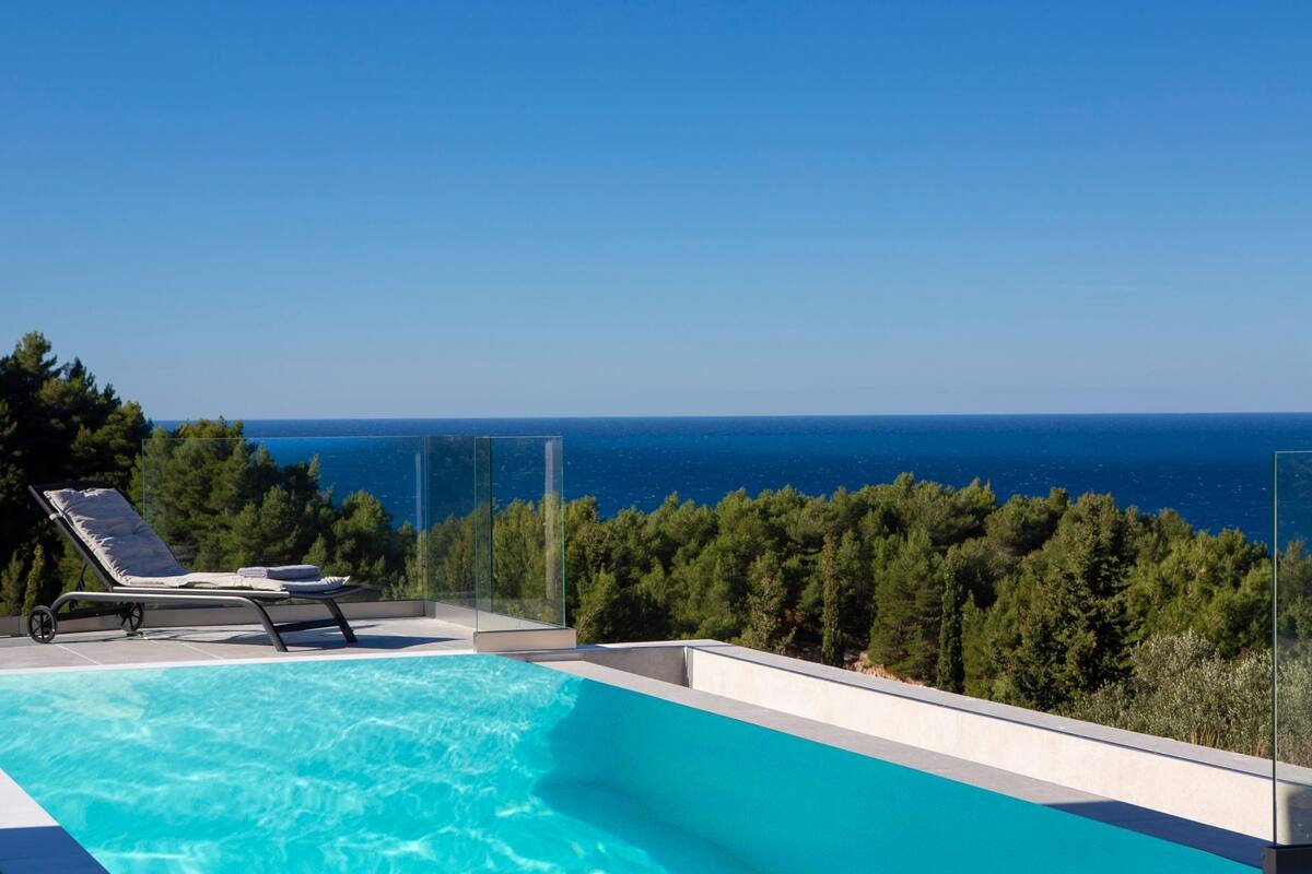 A modern infinity pool with a clear view of the sea and a row of trees in the background.