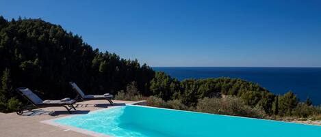 A swimming pool with two sun loungers, surrounded by a forested hillside and overlooking a body of water.