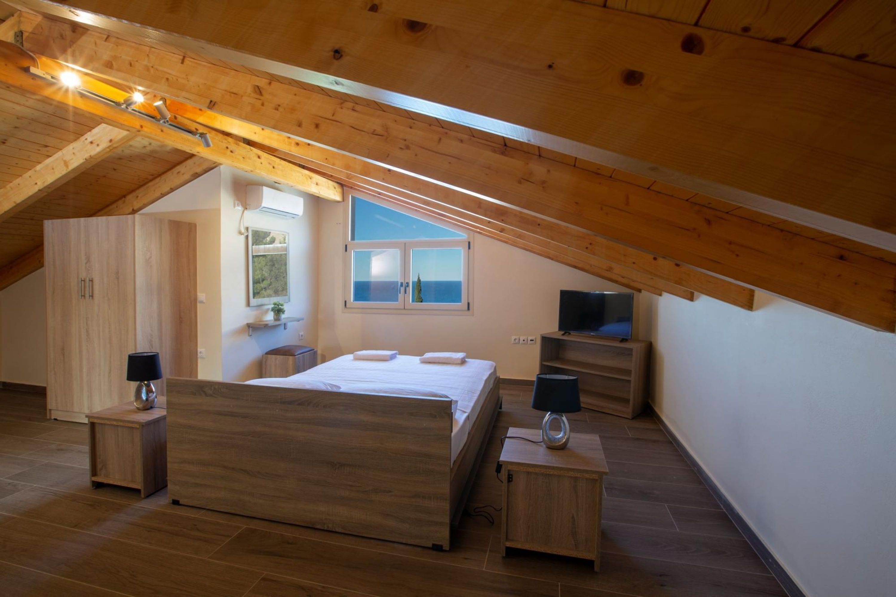 A bedroom with a wooden ceiling, a bed, bedside tables, a TV, and a window with a view.