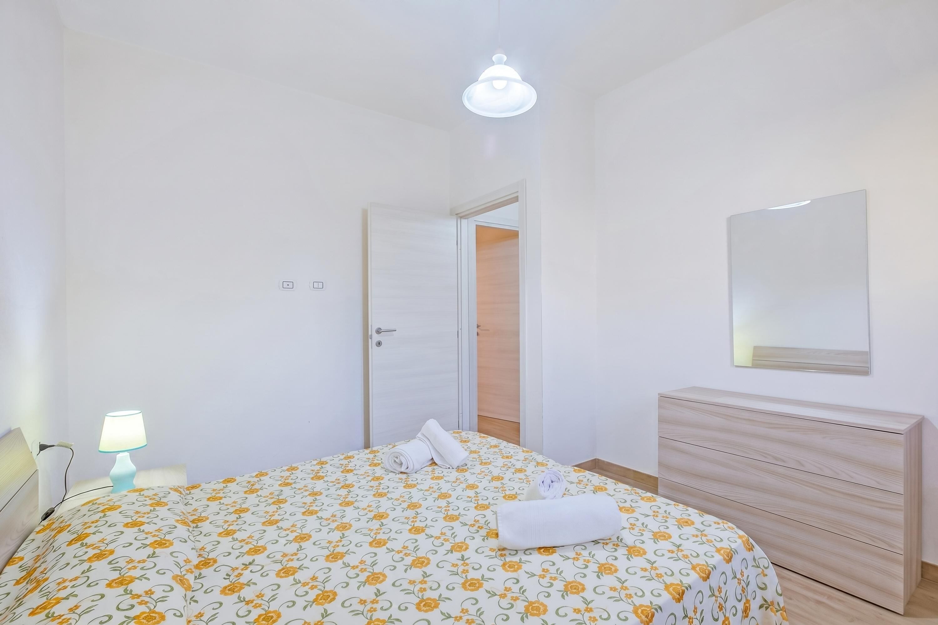 Une chambre à coucher comprenant un lit, une lampe de chevet, une commode, un miroir et une porte donnant sur une autre pièce.