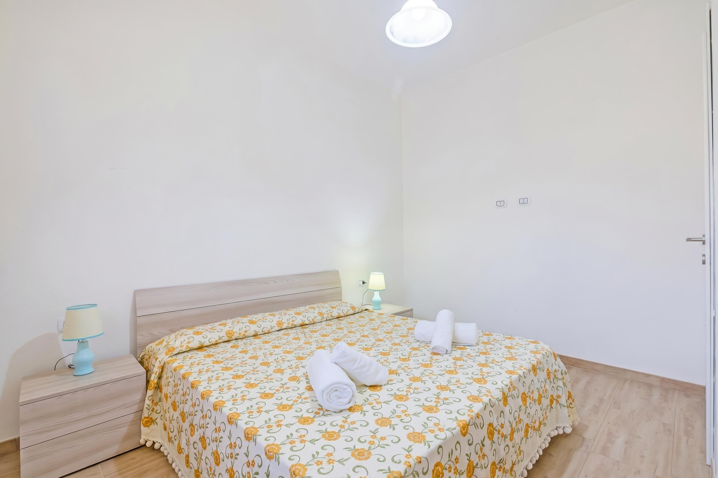 Un letto rifatto con lenzuola a motivi floreali, una lampada da comodino e un asciugamano piegato sul tavolino.