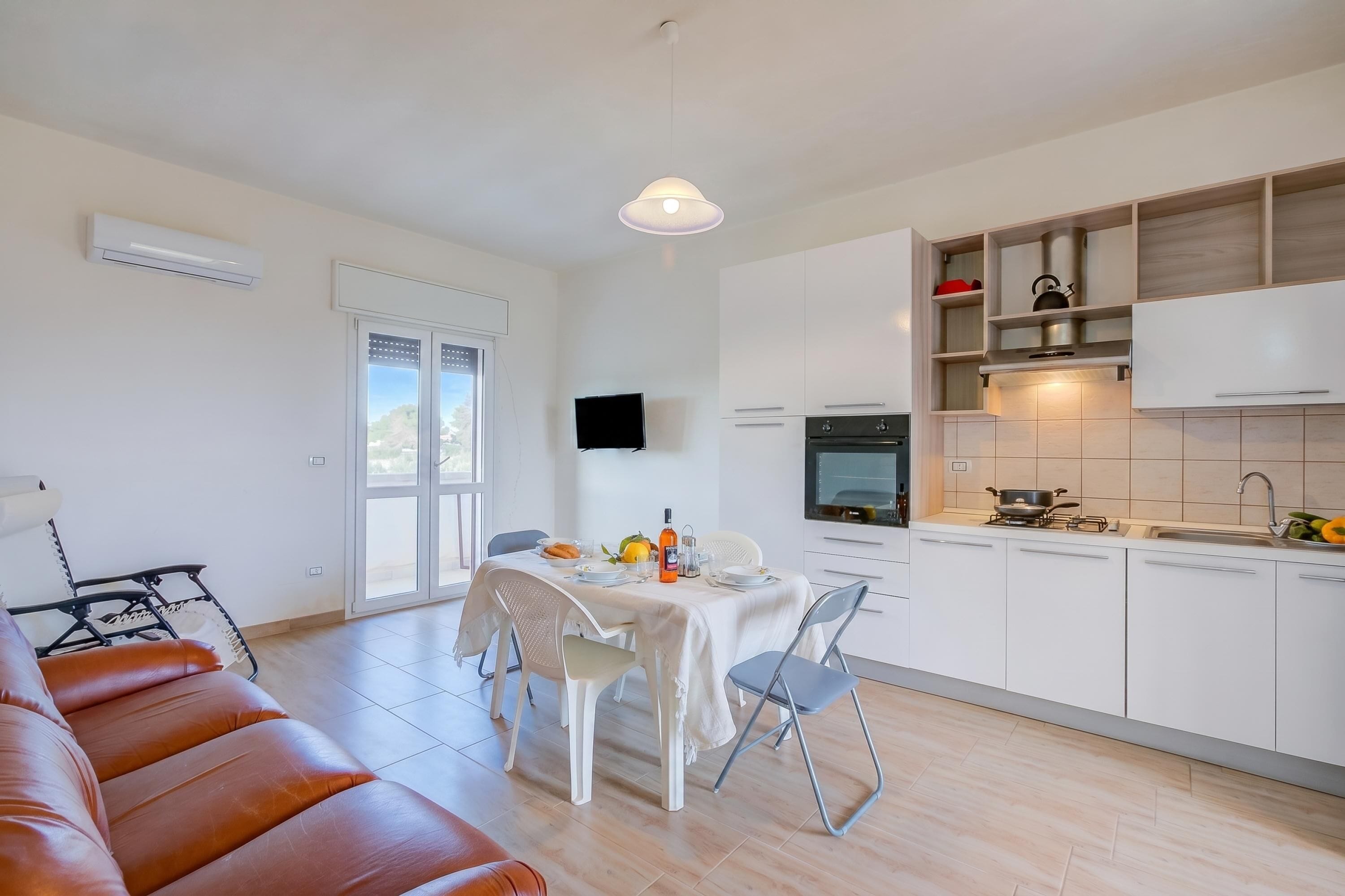 Een moderne keuken met witte kasten, een eettafel met stoelen, een bank en een televisie aan de muur.