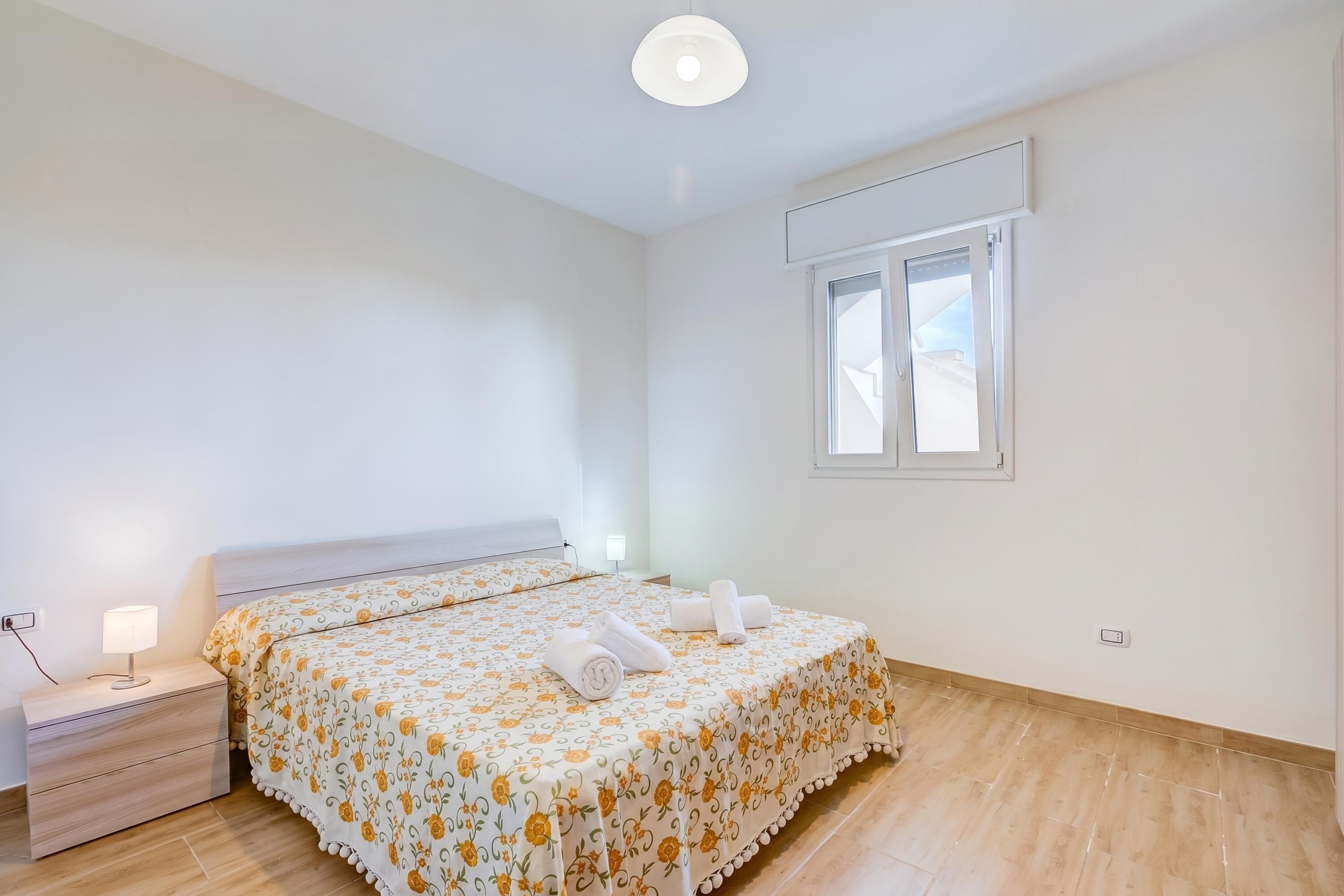 A bedroom with a bed, bedside table, lamp, window, and a view of the sky.
