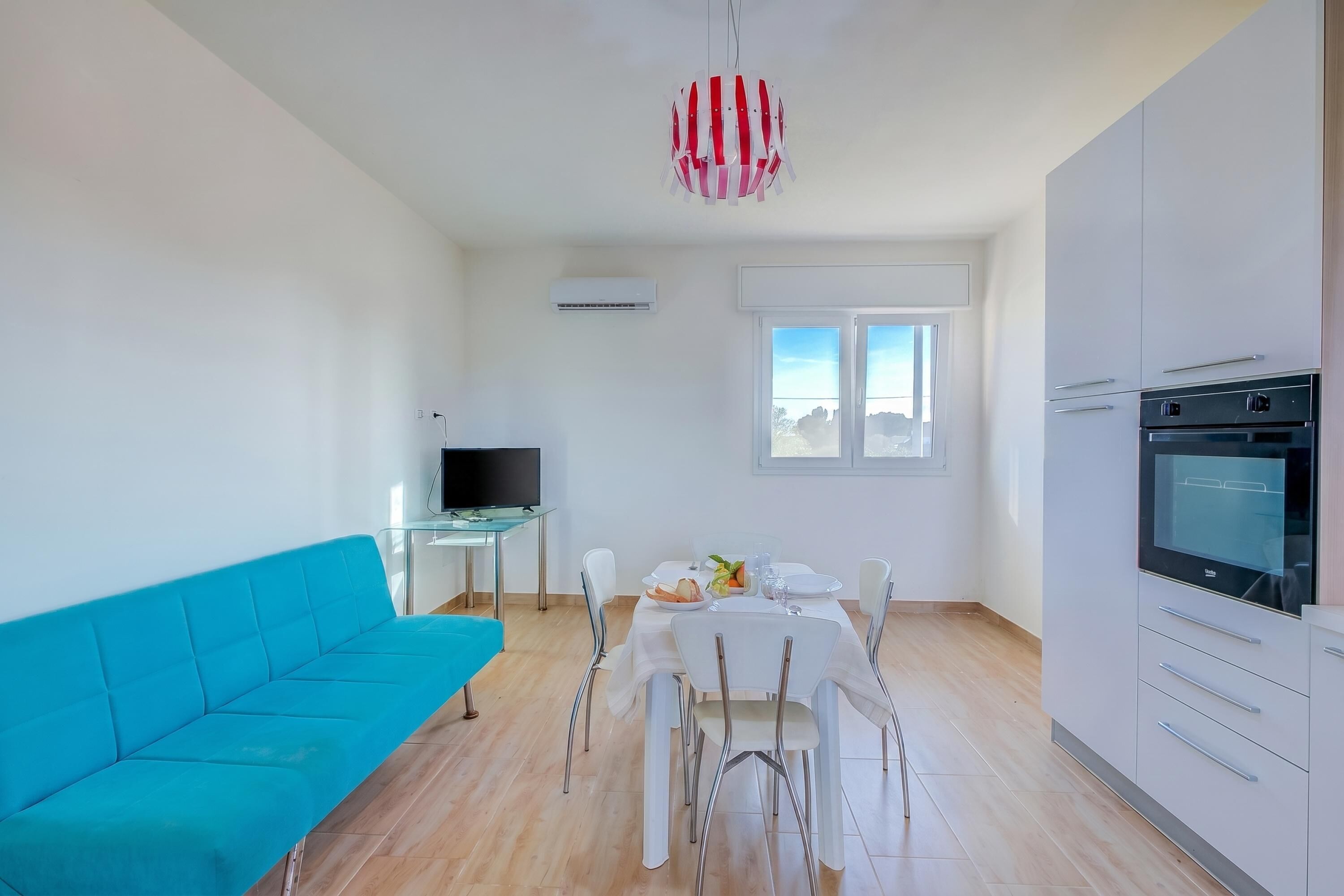 A modern living room with a blue sofa, a dining table with chairs, a TV, and a kitchen area with cabinets and an oven.