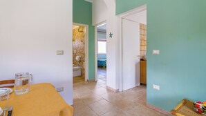 A dining area with a table set for two, a kitchen visible through an open doorway, and a bathroom with a tiled wall. - Villetta Belvedere 400 Meters From Sea (Porto Cesareo)