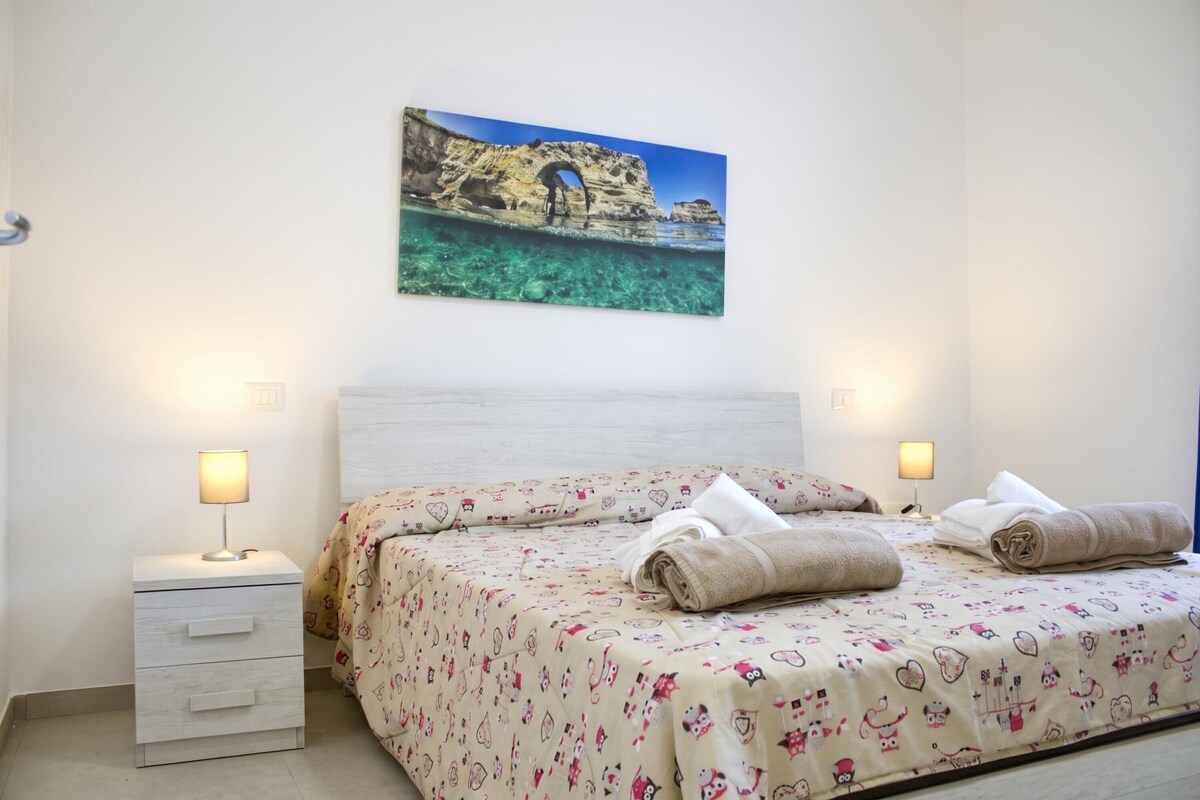 A bedroom with a bed, two bedside lamps, a nightstand, and a wall-mounted painting of a coastal scene.