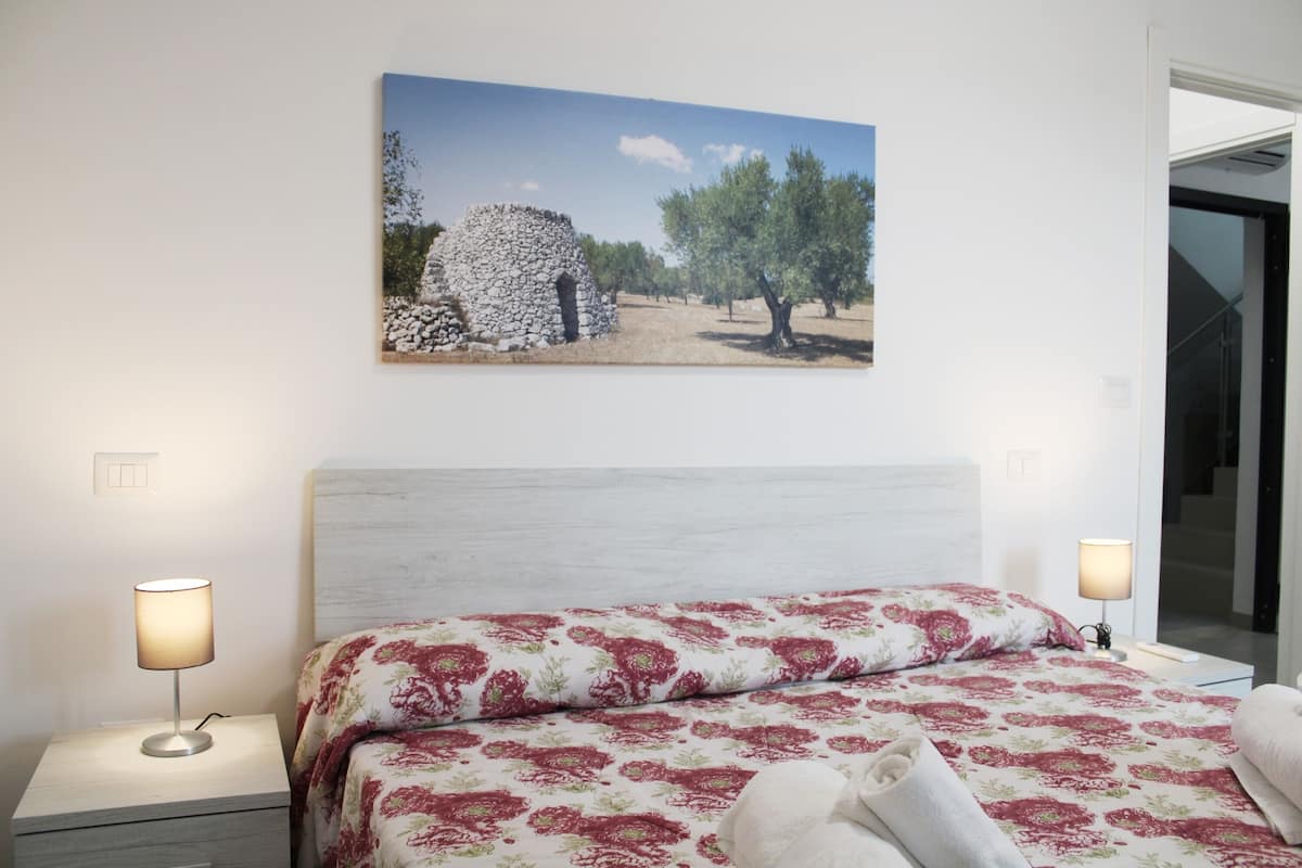 Une chambre à coucher comprenant un lit, deux lampes de chevet, un tableau représentant une structure en pierre et des arbres, et une vue sur un escalier.