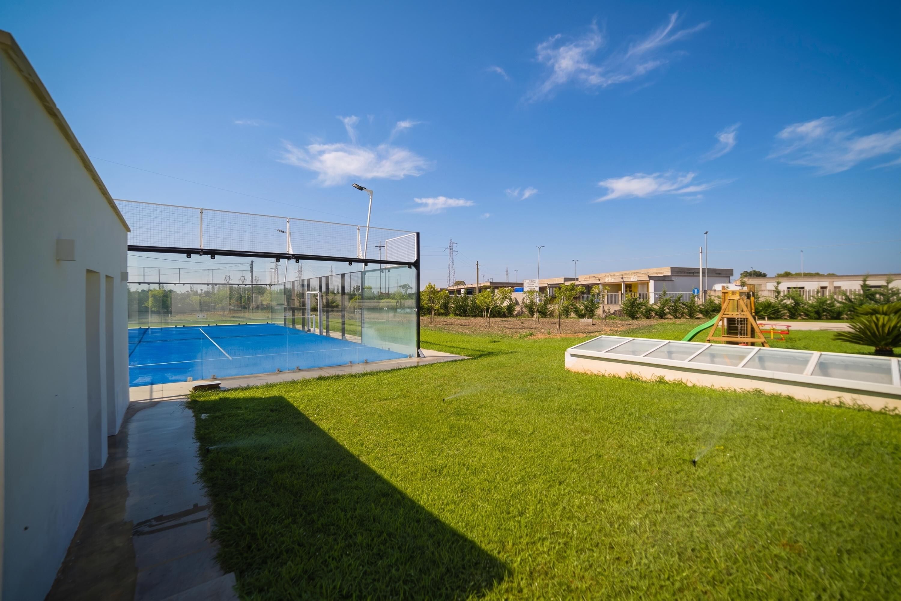 A residential area with a swimming pool, a fenced tennis court, and a grassy lawn.