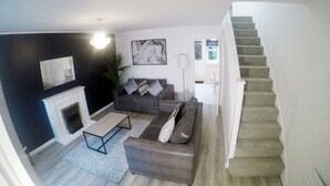 A modern living room with a fireplace, a grey sofa, a coffee table, and a staircase. - Gorgeous 3 bed in Bristol (Bradley Stoke)