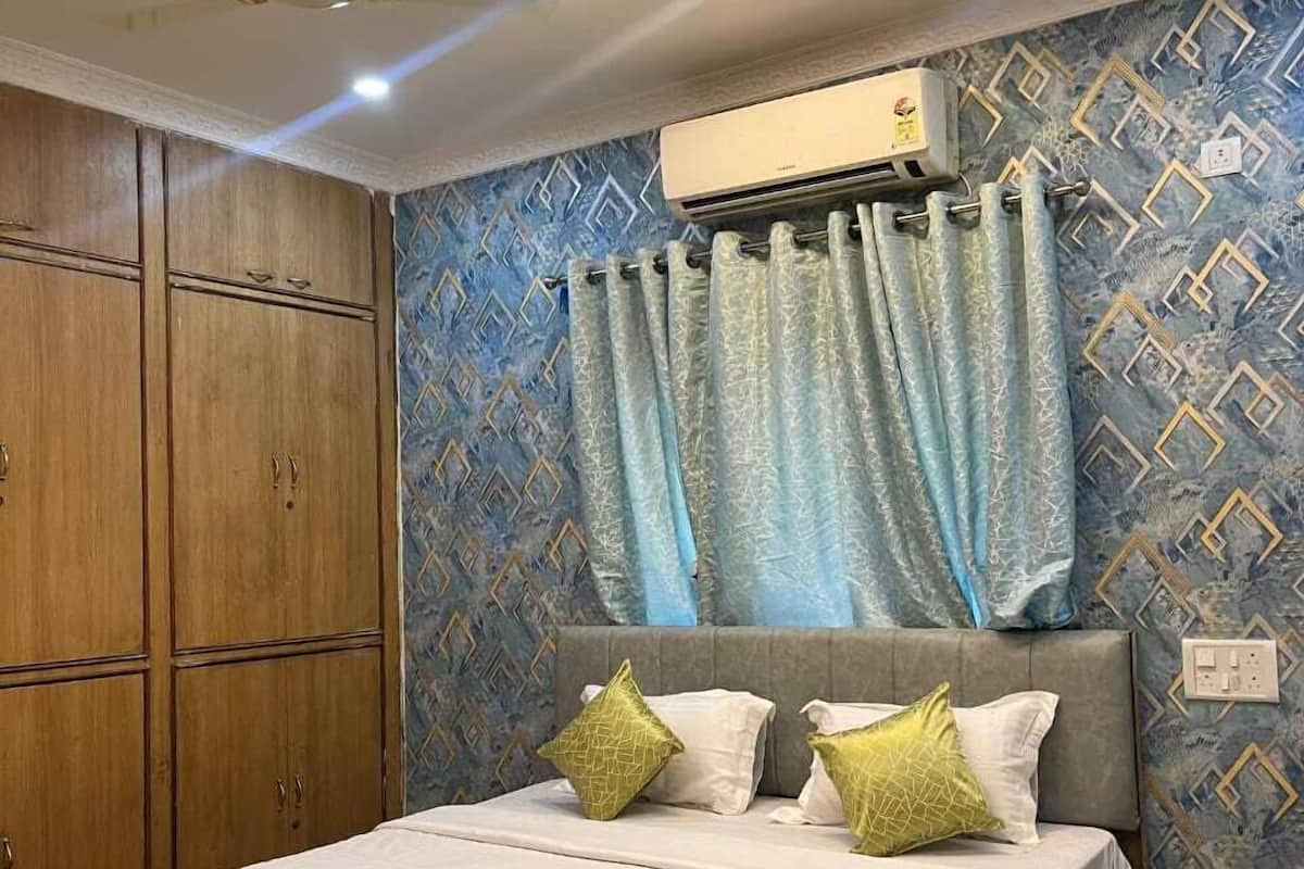 A bedroom with a bed, curtains, an air conditioning unit, and patterned wallpaper.