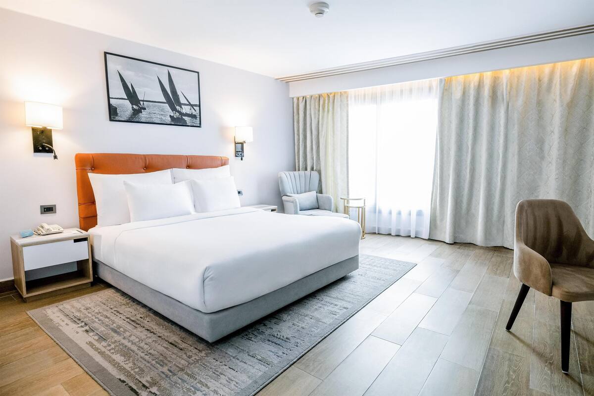 Una habitación de hotel moderna con una cama grande, una mesita de noche, una silla y un ventanal con cortinas.
