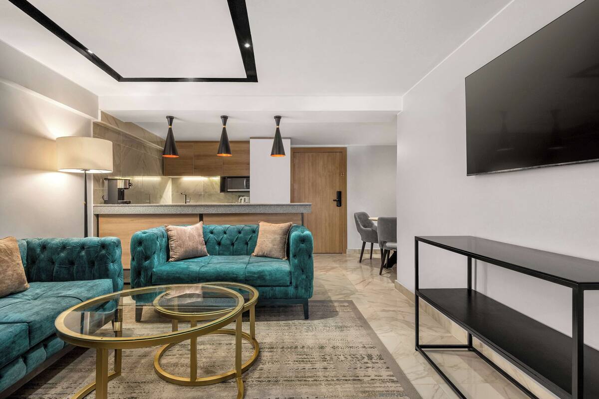 Una sala de estar moderna con un sofá color turquesa, una mesa de centro de vidrio, un televisor de pantalla plana y una cocina al fondo.