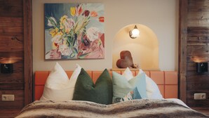 A bedroom with a wooden headboard, a painting of flowers, a mirror with a light, and a bed with a striped blanket. - Apartment Aschbach - cosy for couples or families with 1-2 children  (Kitzbuhel)