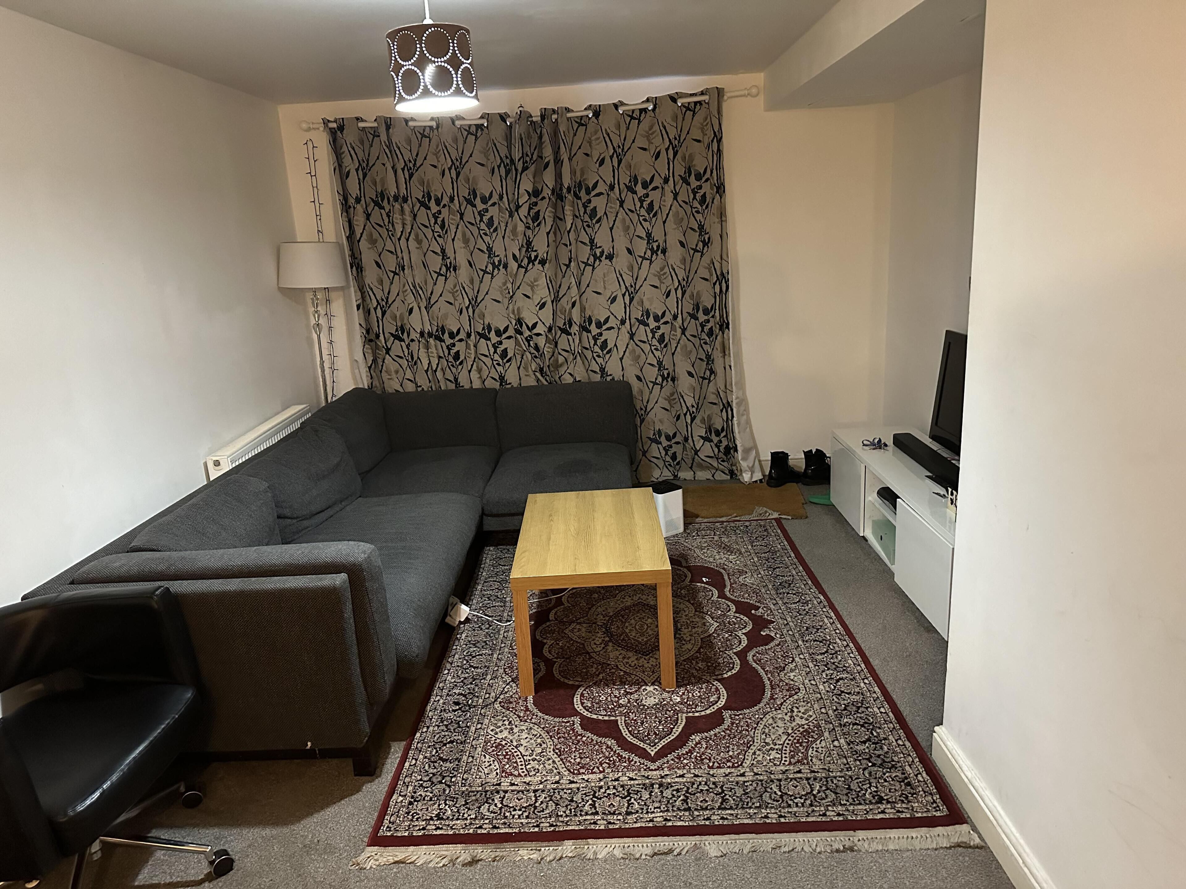 Ein Wohnzimmer mit grauem Sofa, einem Holztisch, einem schwarzen Bürostuhl, einem gemusterten Teppich, einem Fernseher und einer weißen Kommode.