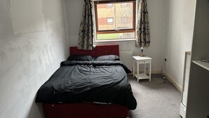 A small, sparsely furnished room with a single bed, a small table, and a window with curtains. - Immaculate 2-bed Apartment in Greenhithe (Greenhithe)