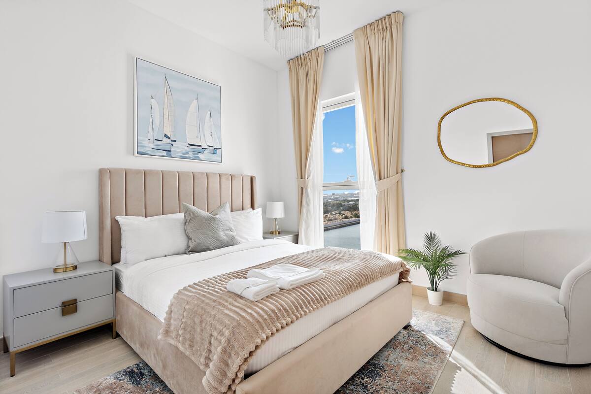 Un dormitorio con cama, mesita de noche, lámpara, planta en maceta y vistas al exterior.