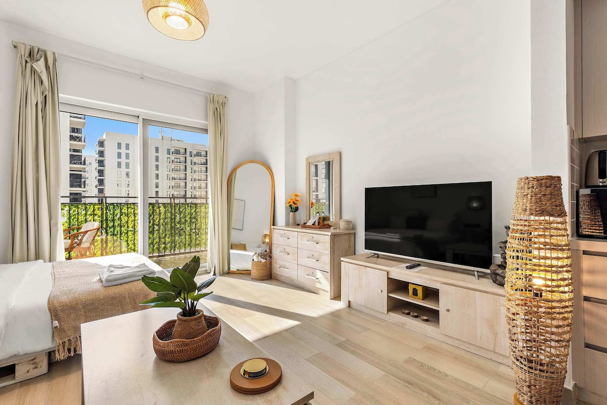 Habitación de hotel moderna con cama, televisor, ventana grande con vistas a edificios y lámpara de mimbre.
