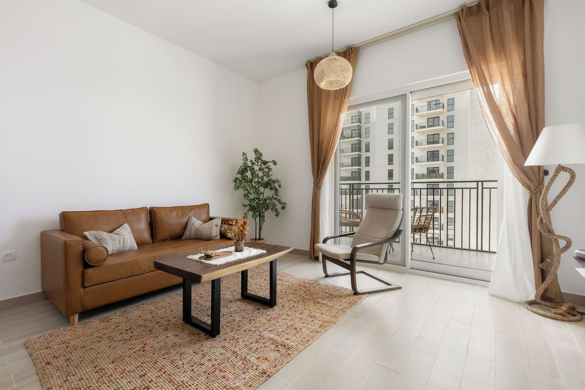 Una sala de estar moderna con un sofá de cuero marrón, una mesa de centro de madera, una silla y un ventanal con vista a un edificio.