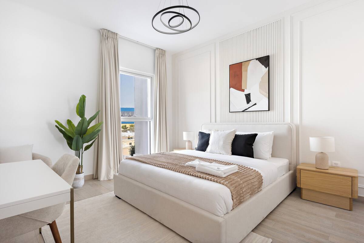 Habitación de hotel moderna con una cama grande, una mesita de noche con lámpara, una planta en maceta y vistas de la ciudad a través de la ventana.