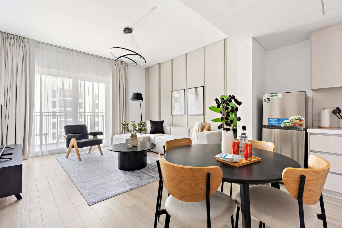 Una sala de estar moderna con una mesa de comedor redonda, sillas, un sofá y una zona de cocina con refrigerador.