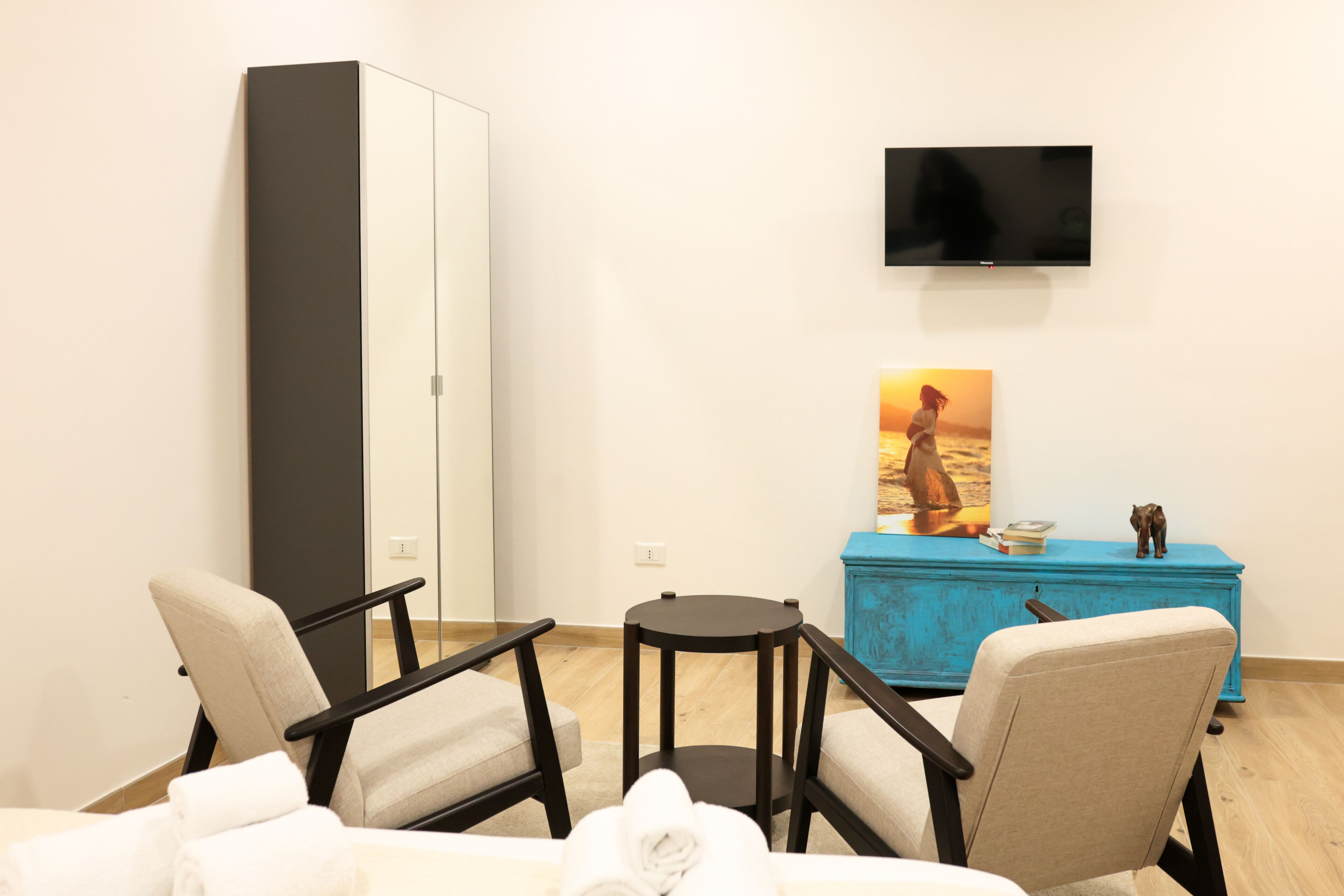 Habitación de hotel con cama, dos sillones, una mesita redonda, televisor montado en la pared y un aparador azul con objetos decorativos.