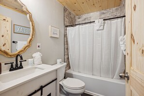 A bathroom with a wooden ceiling, a white bathtub with a shower curtain, a white toilet, and a vanity with a mirror and towel rack. - Majestic Valley View 3 Bedroom Cabin by RedAwning (Sevierville)