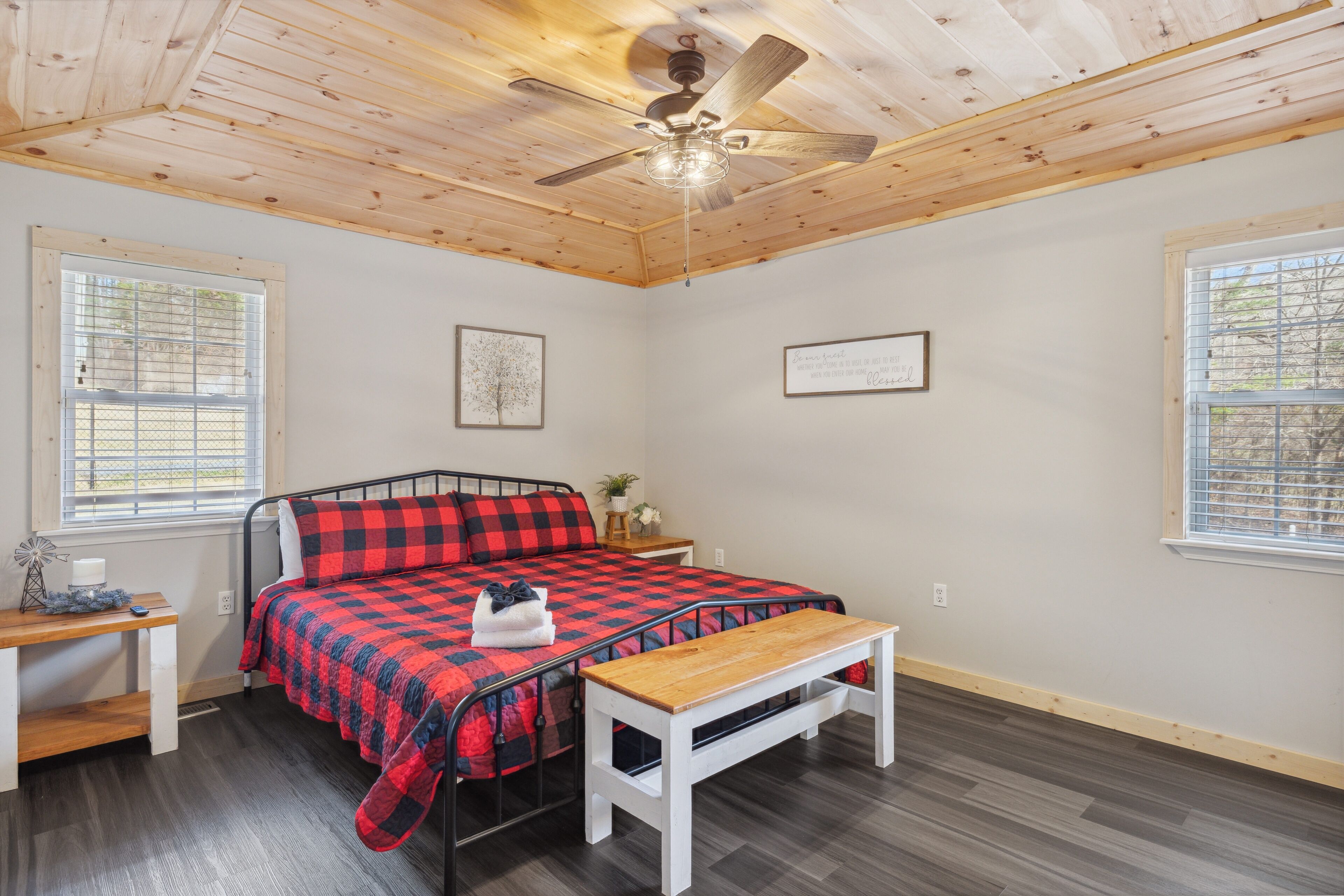 A bedroom with a bed, a wooden bench, a ceiling fan, and two windows with blinds.