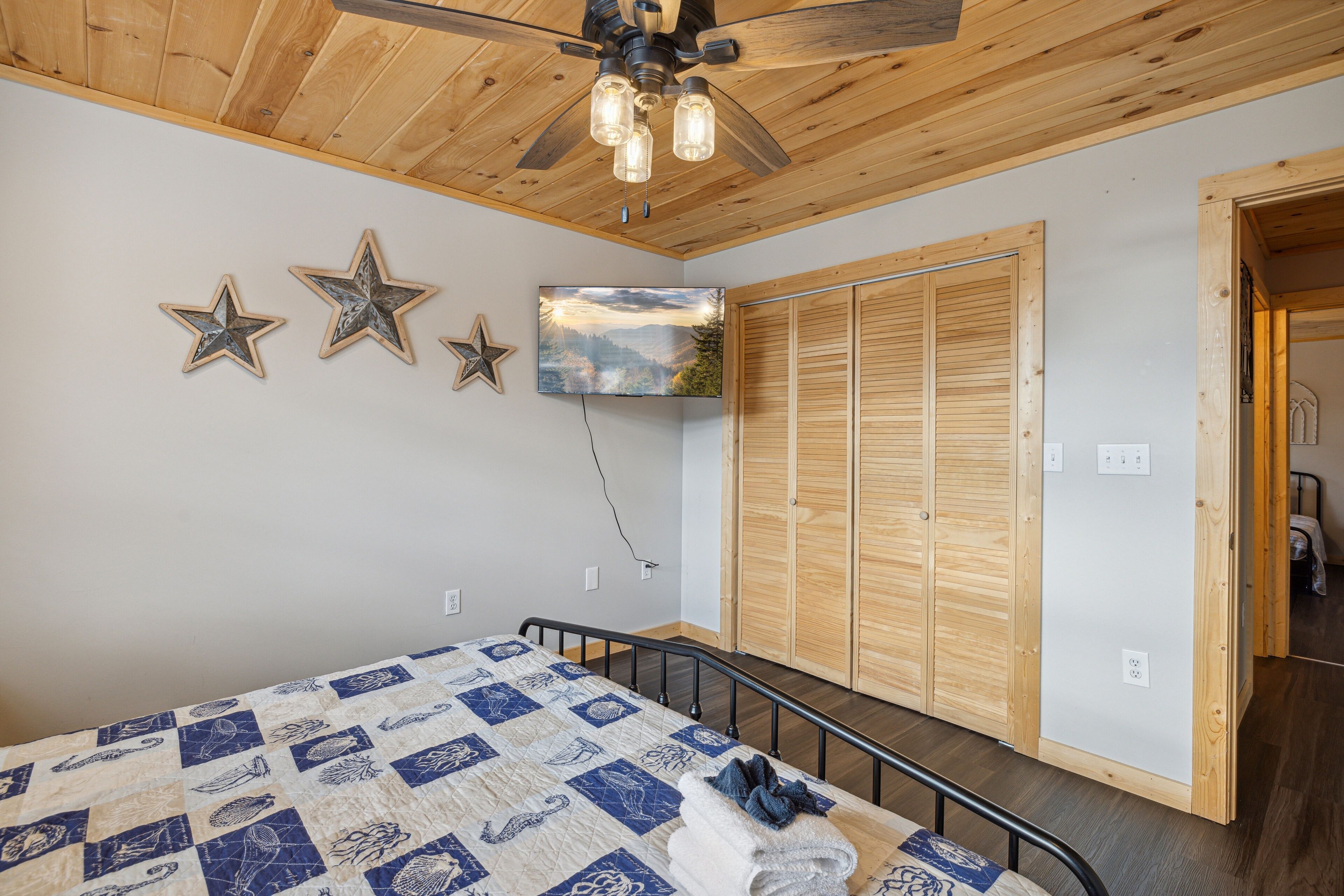 Une chambre avec un plafond en bois, un lit recouvert d’une courtepointe, un ventilateur de plafond, un téléviseur fixé au mur et des armoires en bois.