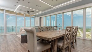 A spacious dining area with a wooden table and chairs, large windows offering a view of the ocean, and a ceiling fan. - Phoenix Gulf Towers 2601 4 Bedroom Condo (Orange Beach)