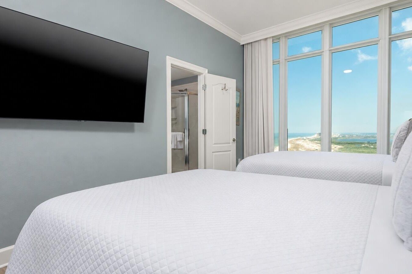 A hotel room with a large bed, a TV mounted on the wall, a door leading to a bathroom, and a view of the beach through large windows.