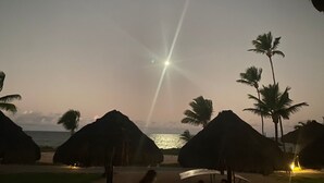 Une scène de plage avec des huttes aux toits de chaume et des palmiers, un ciel dégagé et l’océan en arrière-plan.