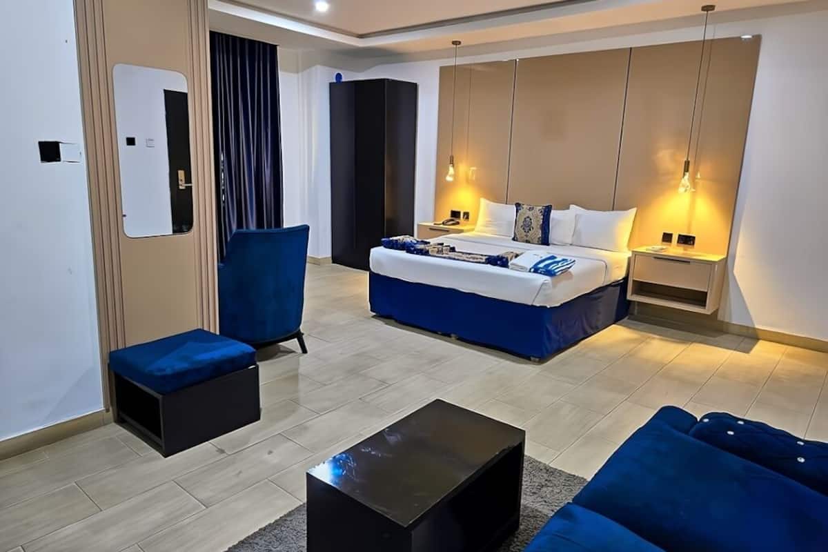 A modern hotel room with a large bed, a sofa, a coffee table, and a desk with a lamp.
