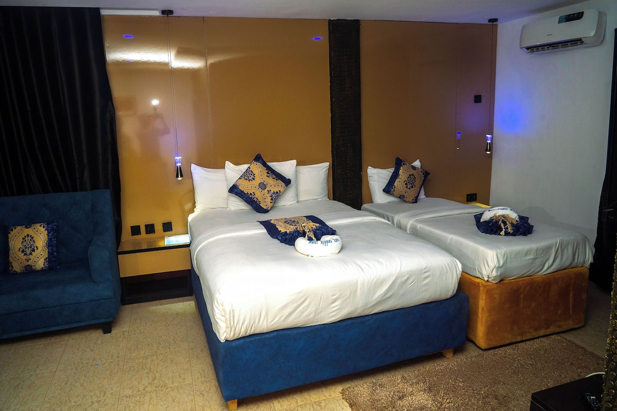 A hotel room with a large bed, a smaller bed, a blue armchair, and a wall-mounted air conditioner.