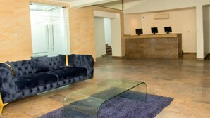 A spacious lobby with a dark blue sofa, a glass coffee table, and a reception desk with three monitors. - Water Ripples Hotel (Lekki)