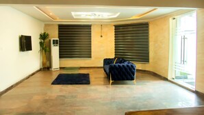 A spacious room with a large window, a dark blue sofa, a small rug, and a wall-mounted TV. - Water Ripples Hotel (Lekki)