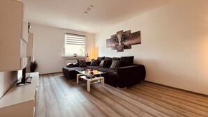Un salon moderne avec un canapé noir, une table basse et une grande fenêtre équipée de stores.
