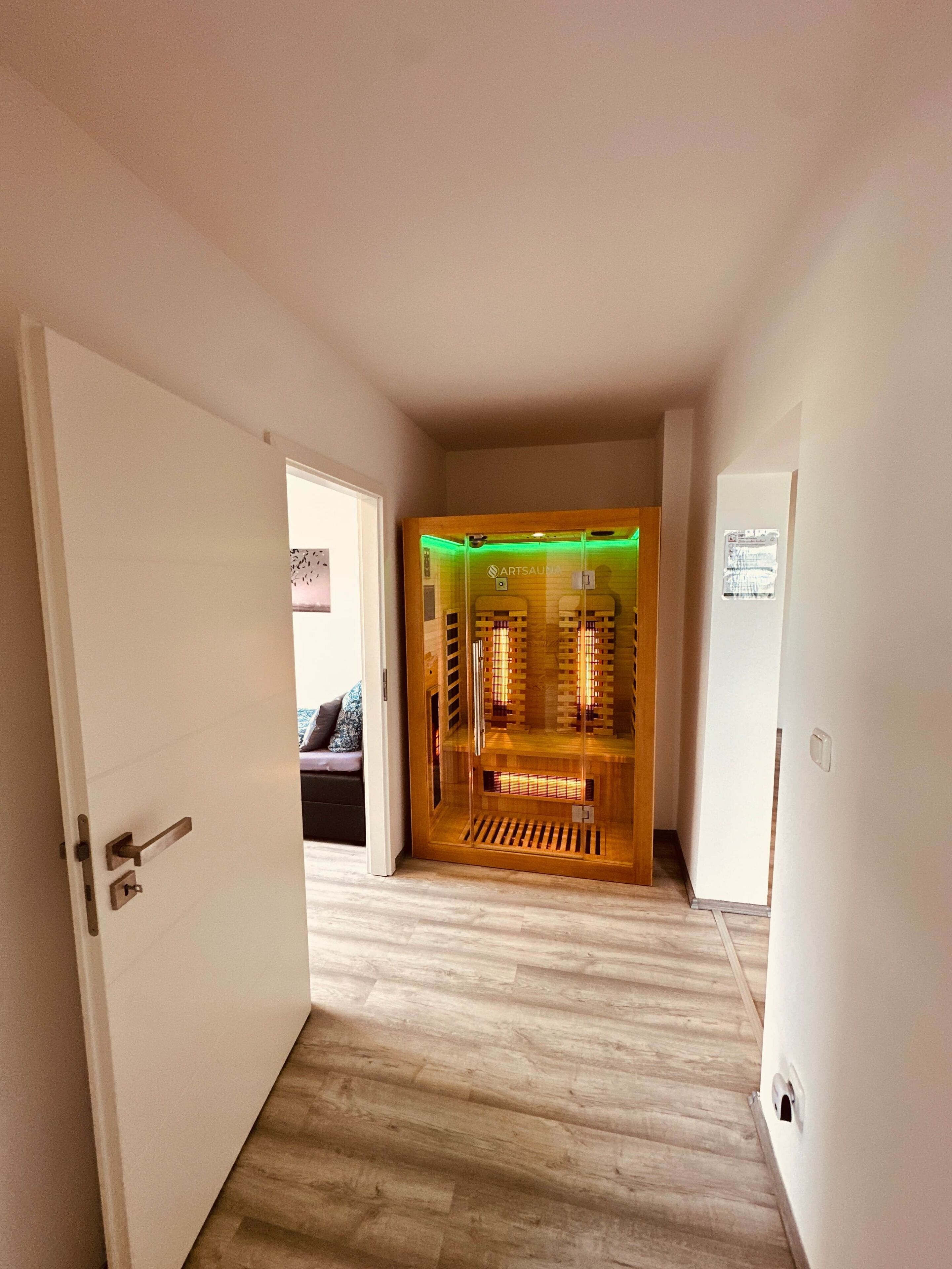A hallway leading to a sauna room with a glass door, wooden floor, and a visible sauna interior.