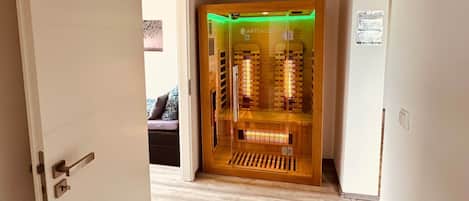 A hallway leading to a sauna room with a glass door, wooden floor, and a visible sauna interior.